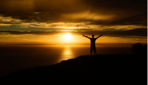 A man reaching his arms out towards the heavens with the sun on the horizon
